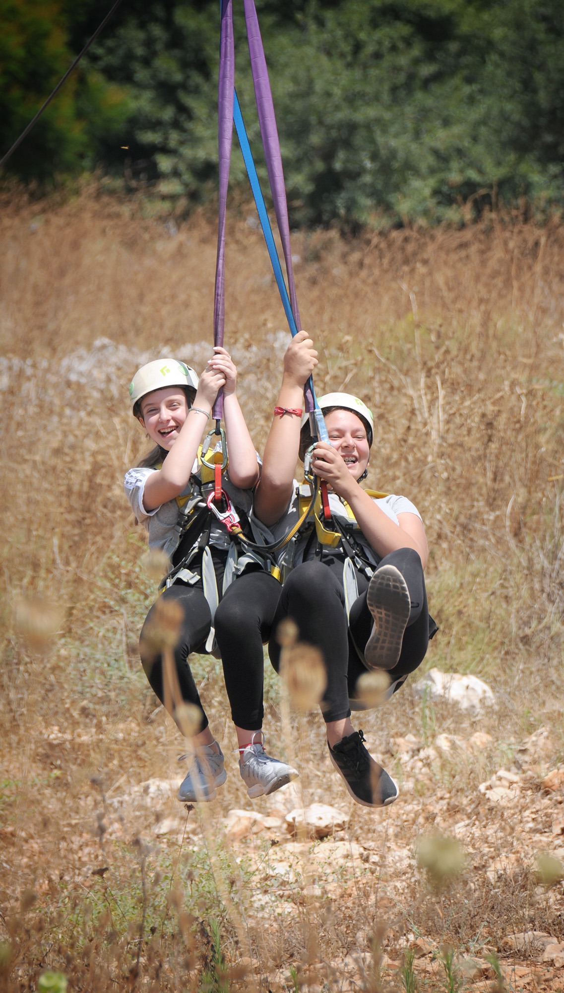 Two campers glide through the air on a tandem zipline at Jordan River Valley, gripping the ropes and laughing as they soar over dry grass and trees. Both wear helmets and harnesses, enjoying the exhilarating outdoor adventure.