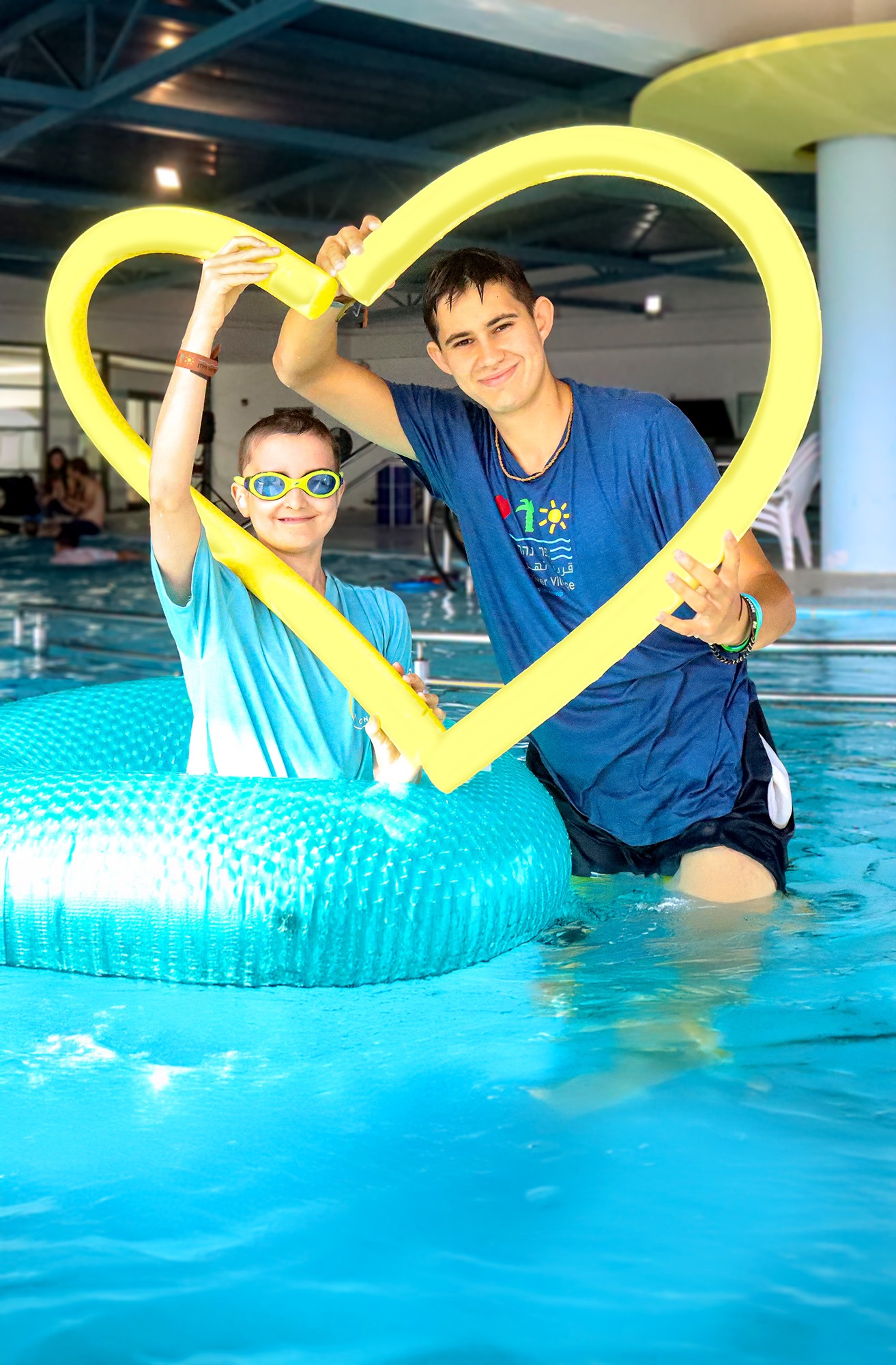 In the Jordan River Valley indoor swimming pool, a young camper in a floatation ring and a counselor hold a yellow pool noodle shaped like a heart. Both are smiling, capturing a moment of connection and joy in the water.