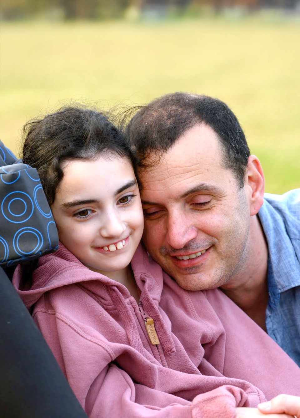 A young camper at Jordan River Valley, dressed in a cozy pink hoodie, sits in a wheelchair while sharing a warm embrace with her smiling father. The father and daughter duo lean their heads together, enjoying a quiet moment outdoors with a blurred grassy field in the background.